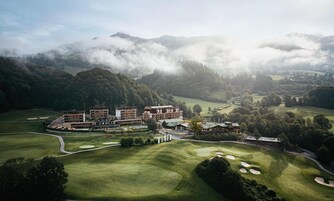 Un campo de golf con un complejo hotelero, rodeado de colinas boscosas y neblina.
