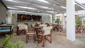 An outdoor patio area with wooden tables and chairs, a bar with a counter and shelves, and a white column structure. - Olga's Pension (Ios)