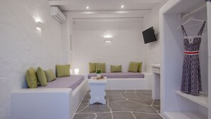 A spa room with a stone floor, white walls, green and patterned pillows, a white bench, a small table with a plant, and a TV mounted on the wall. - Olga's Pension (Ios)