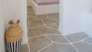 A narrow hallway leading to a bedroom with a stone floor and a bed with a purple blanket. - Olga's Pension (Ios)