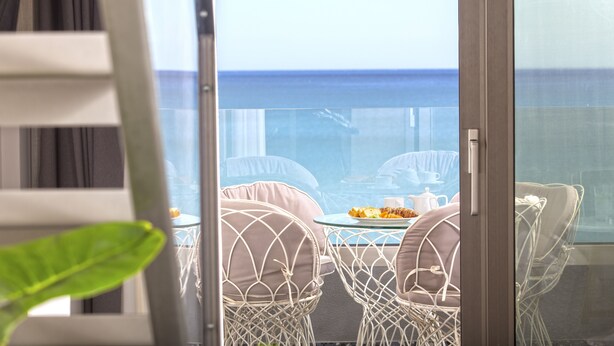 A balcony with a glass door, white wicker chairs, and a table with a plate of food. -  Pearl SeaBreeze Suites (Rethymno)