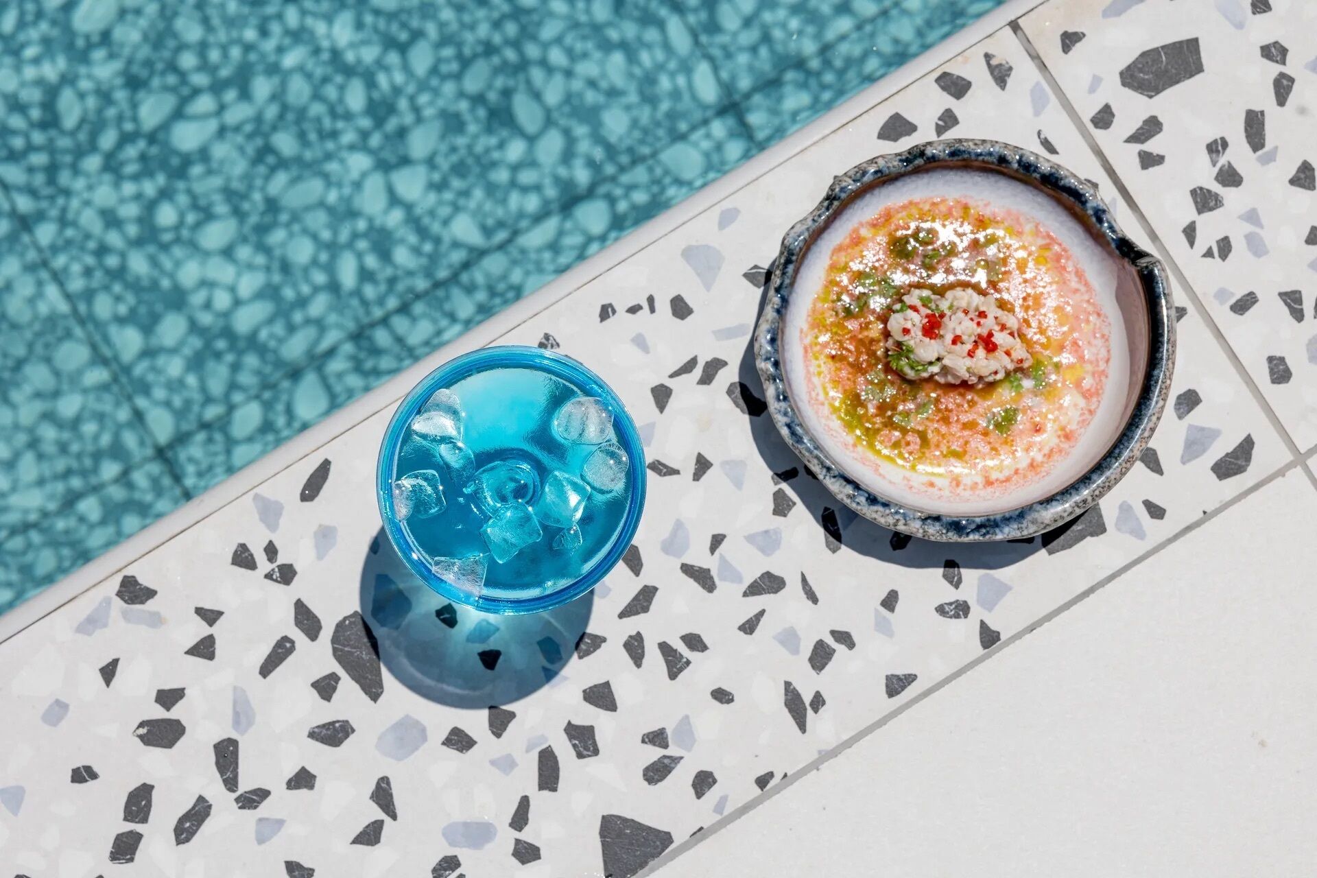 Une boisson bleue avec des glaçons et un bol de soupe garni, le tout posé sur une surface carrelée au bord d’une piscine.