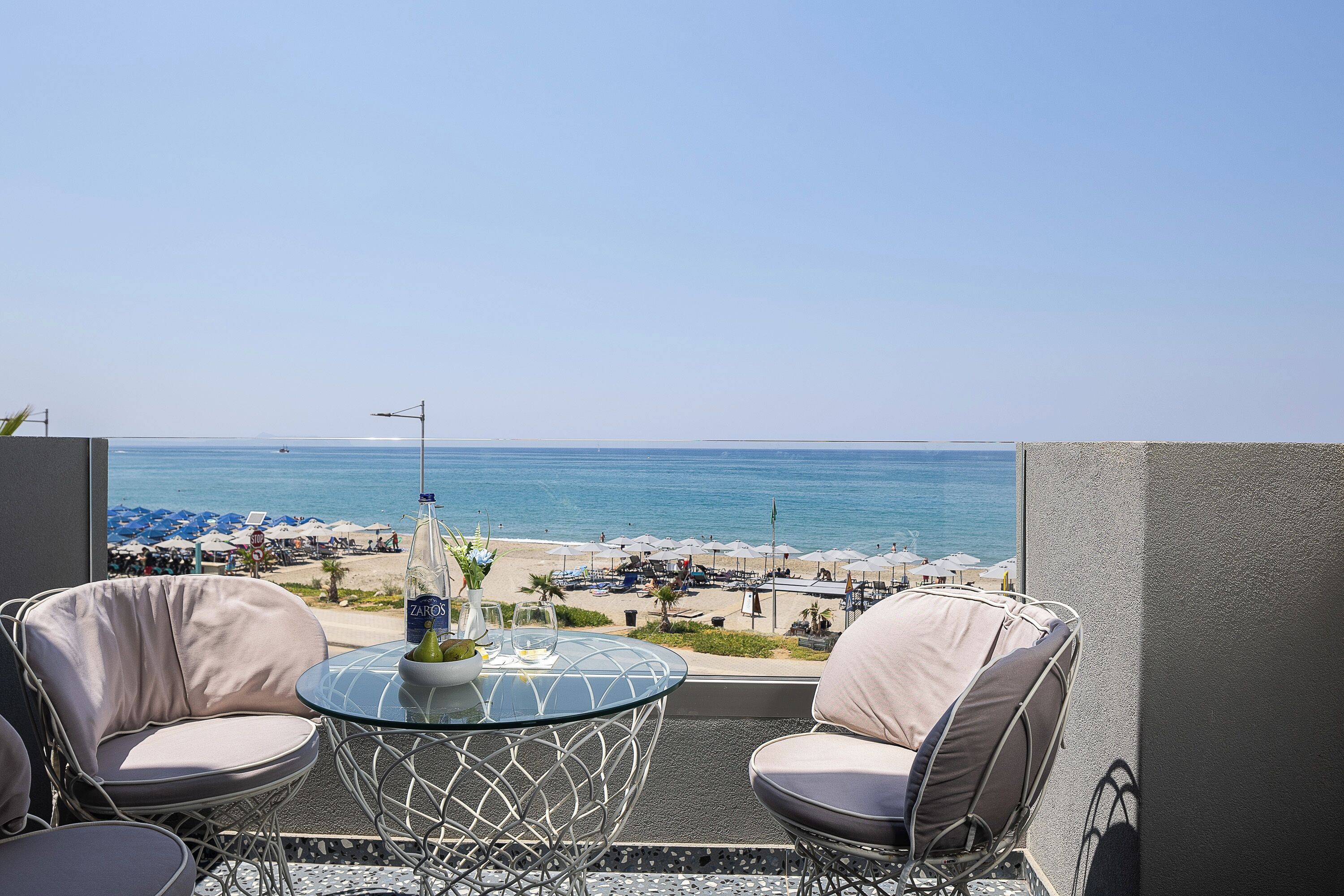A balcony with two cushioned chairs, a glass table, and a bottle of ZAROS. The view includes a beach with umbrellas and a clear sky.