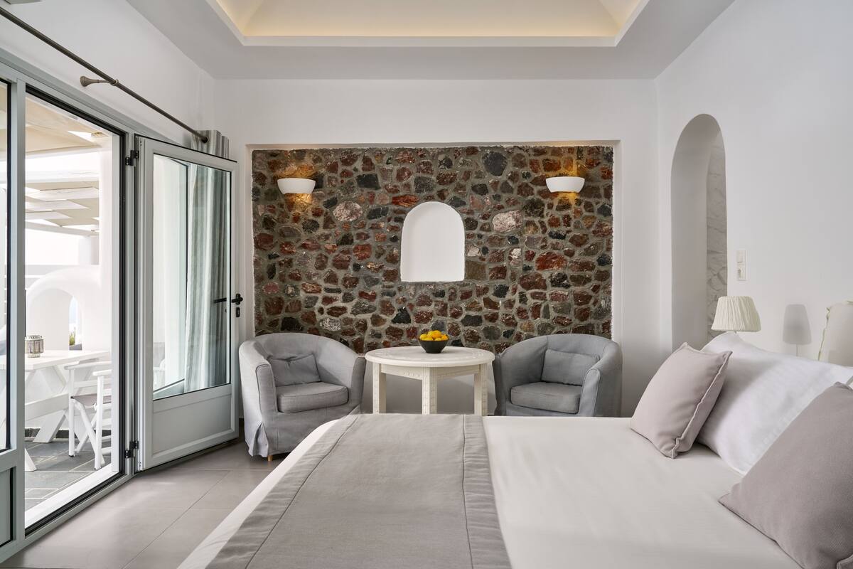 Quarto de hotel moderno com parede de pedra, cama com roupa de cama cinzenta, duas poltronas cinzas, uma mesa redonda pequena com uma cesta de frutas e uma grande porta em arco.