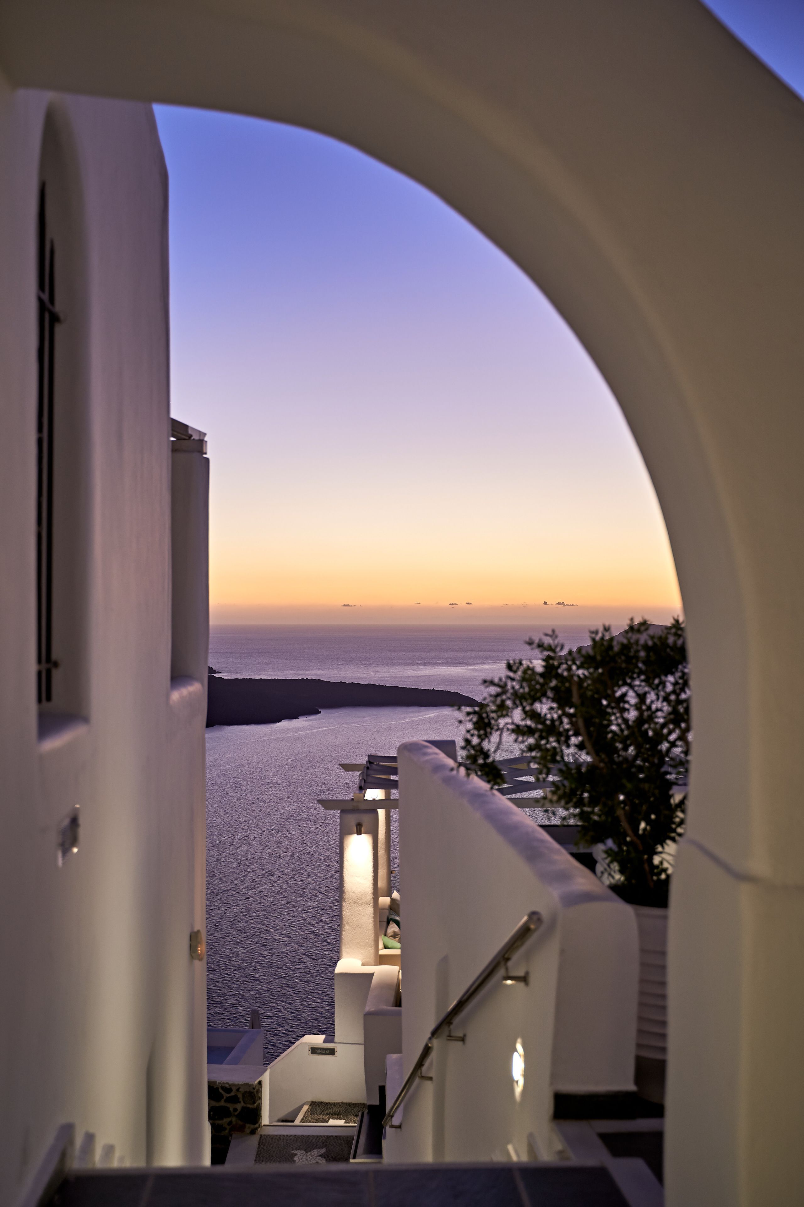 A view from a balcony overlooking the sea at sunset, with a white building and a staircase leading down.