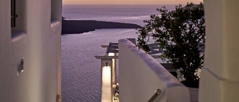 A view from a balcony overlooking the sea at sunset, with a white building and a staircase leading down.