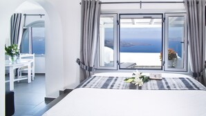 Un dormitorio con una cama grande, una zona de comedor con mesa y sillas, y vista al mar a través de ventanas abiertas.