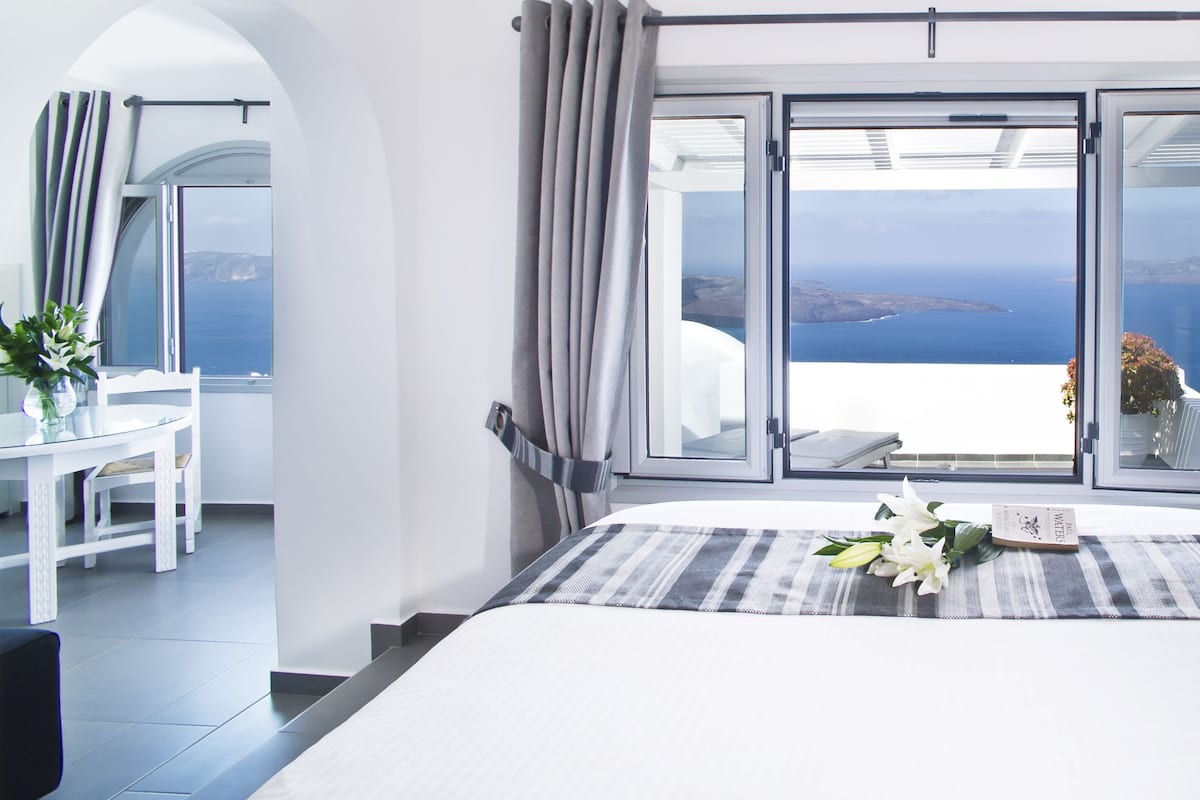 Quarto com uma cama grande, área de jantar com mesa e cadeiras, e vista para o mar pelas janelas abertas.