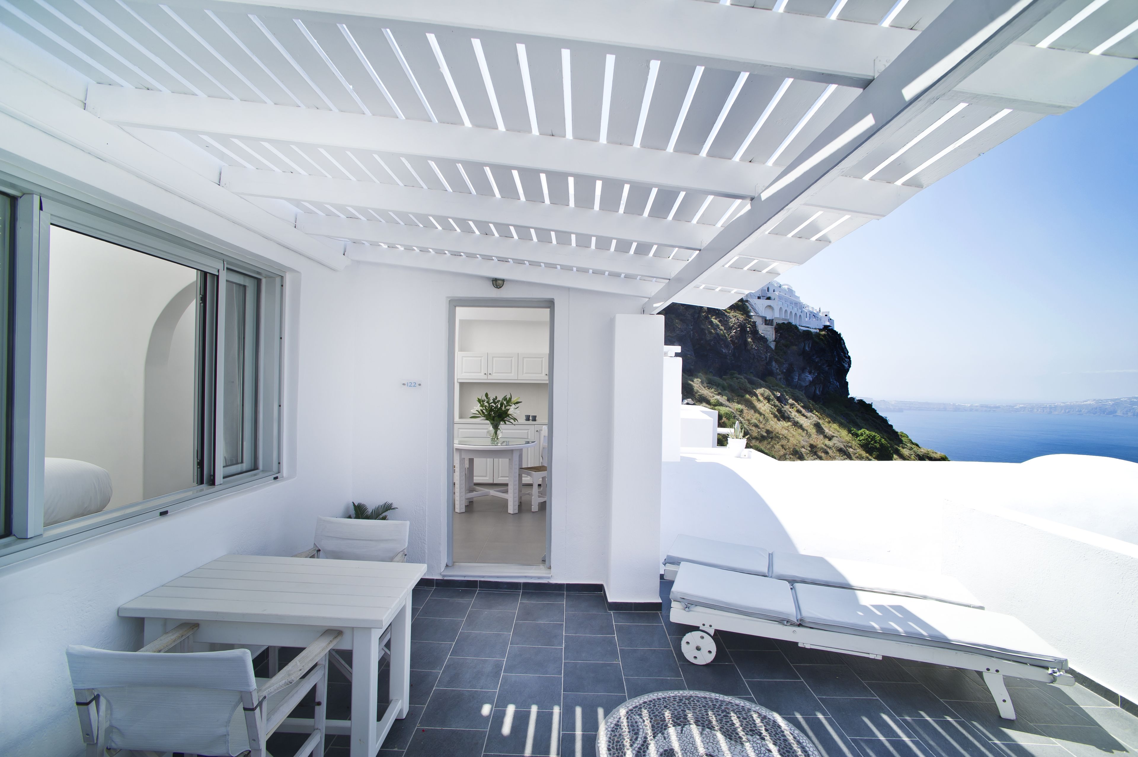 A white room with a table and chairs, a small kitchen area, and a view of the sea and a mountain.