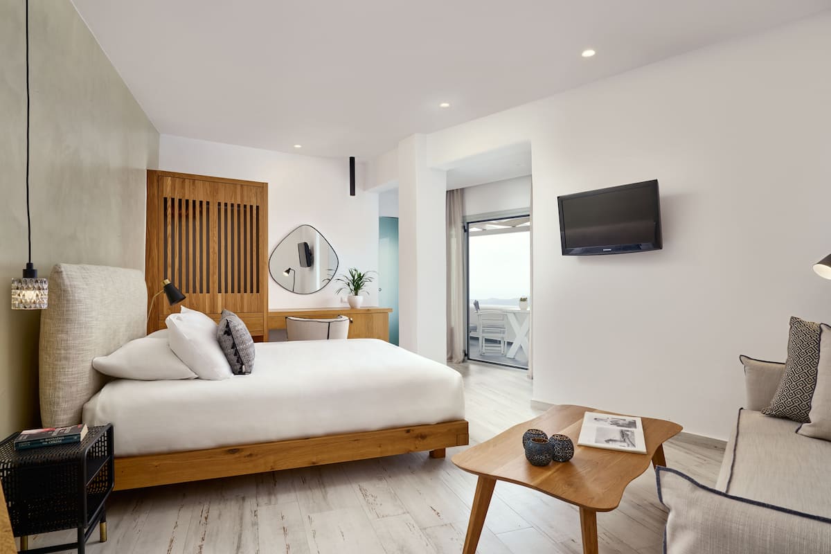 Quarto de hotel moderno com cama de madeira, uma pequena mesa de trabalho, televisão e vista para o exterior.