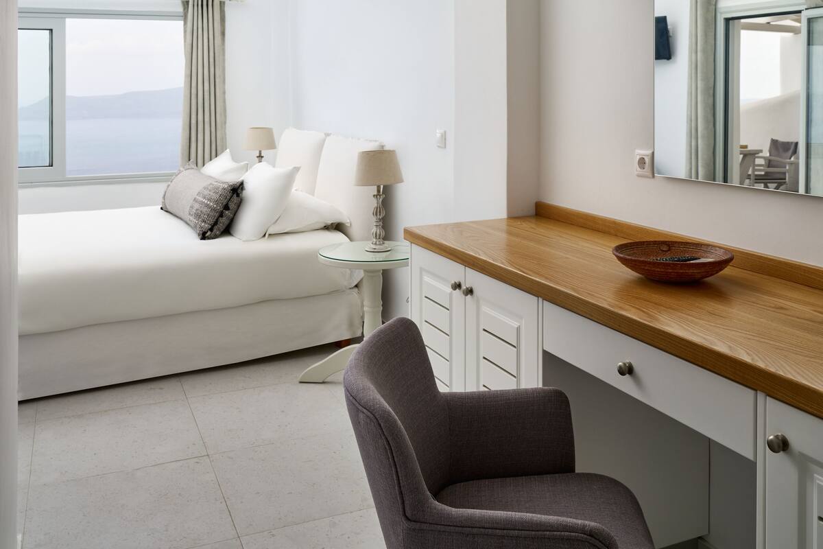 Quarto moderno com uma cama grande, uma escrivaninha de madeira, uma cadeira e vista para o mar pela janela.