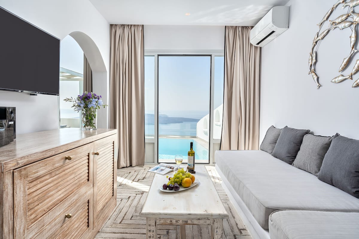 Uma sala de estar moderna com um sofá, um armário de madeira, uma mesa de centro com frutas e vinho, e uma janela grande com vista para o mar.
