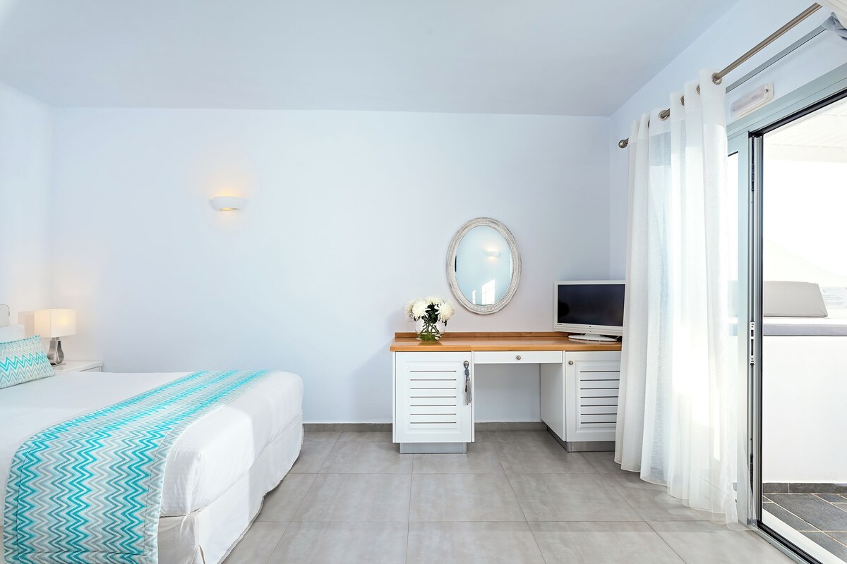 A modern hotel room with a bed, a desk with a TV, a mirror, and a sliding glass door.