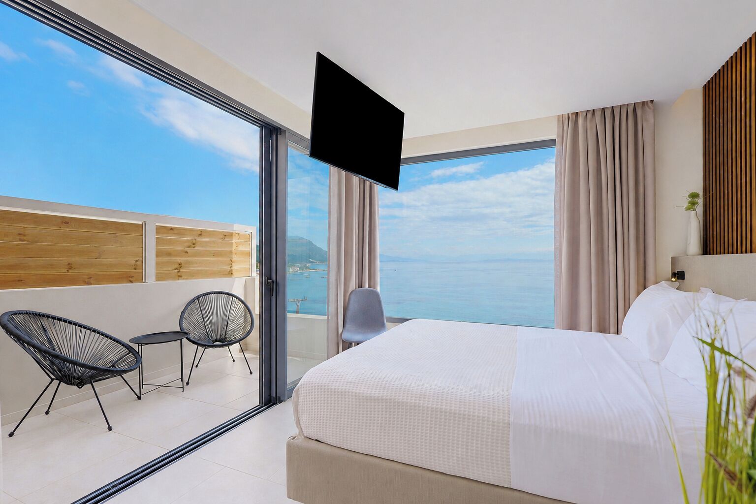 Una habitación de hotel con cama, televisión, balcón con sillas y vista al mar.
