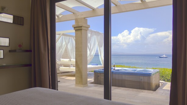 A hotel room with a large window overlooking a pool area, a hot tub, and a view of the sea. - Palms and Spas Corfu Villas Retreat (Corfu)