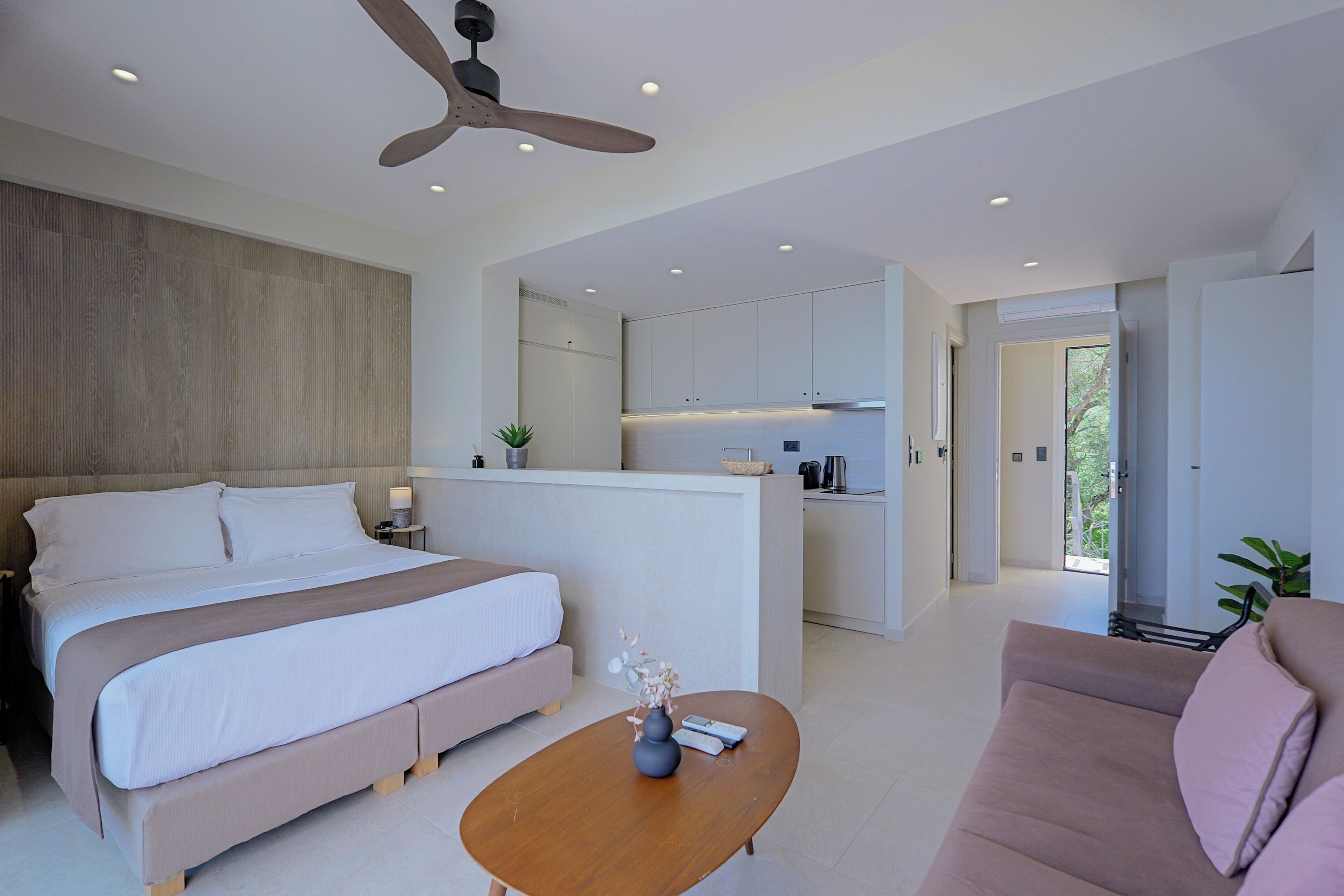 A modern hotel room with a large bed, a sofa, a wooden coffee table, a ceiling fan, and a kitchenette.