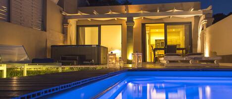 Une piscine dont le bord est illuminé de lumière bleue, un espace extérieur couvert avec des fauteuils de détente et une porte vitrée donnant sur un espace intérieur.