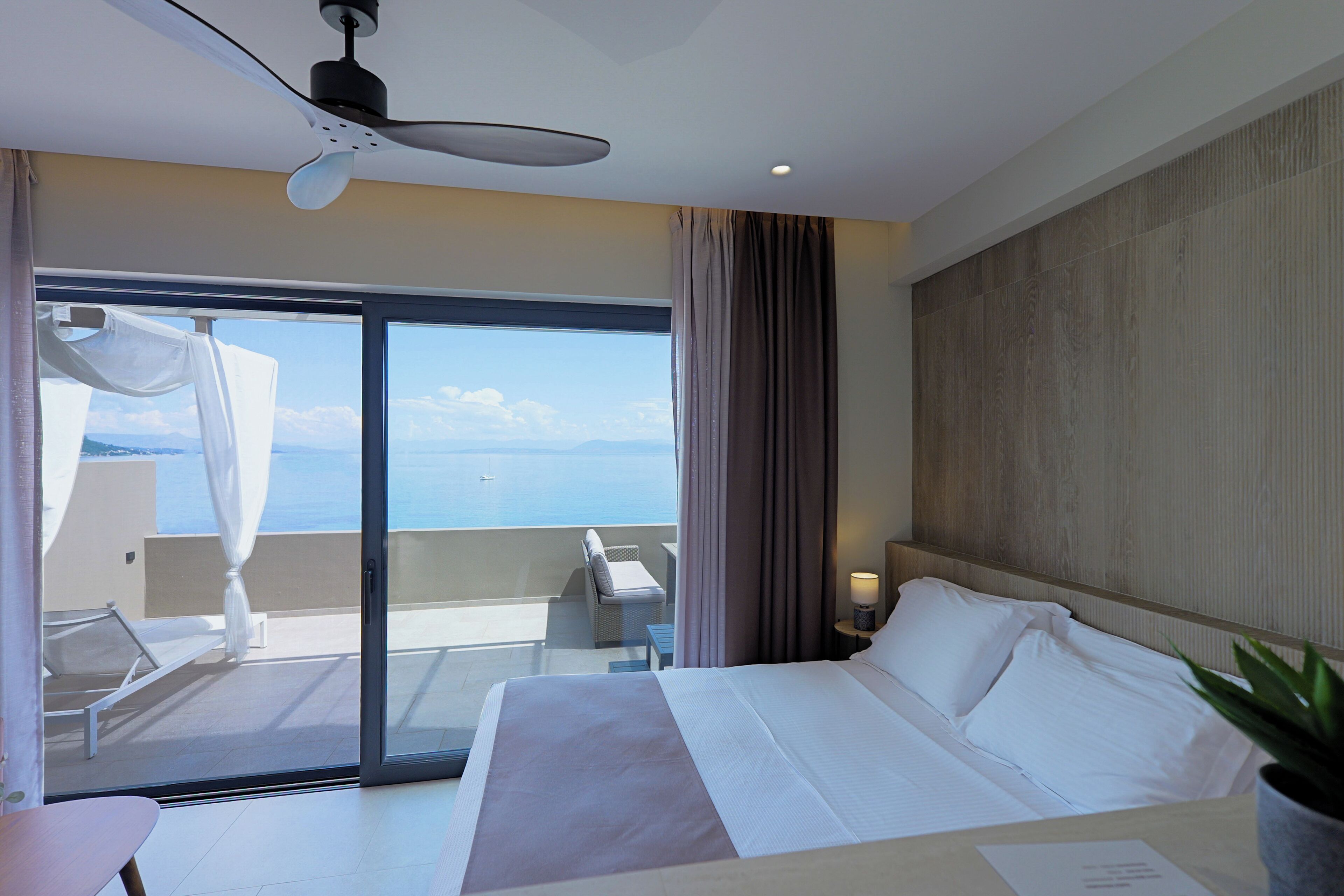 Habitación de hotel con cama, ventilador de techo, vista al mar, balcón con una silla y mesita de noche con lámpara.