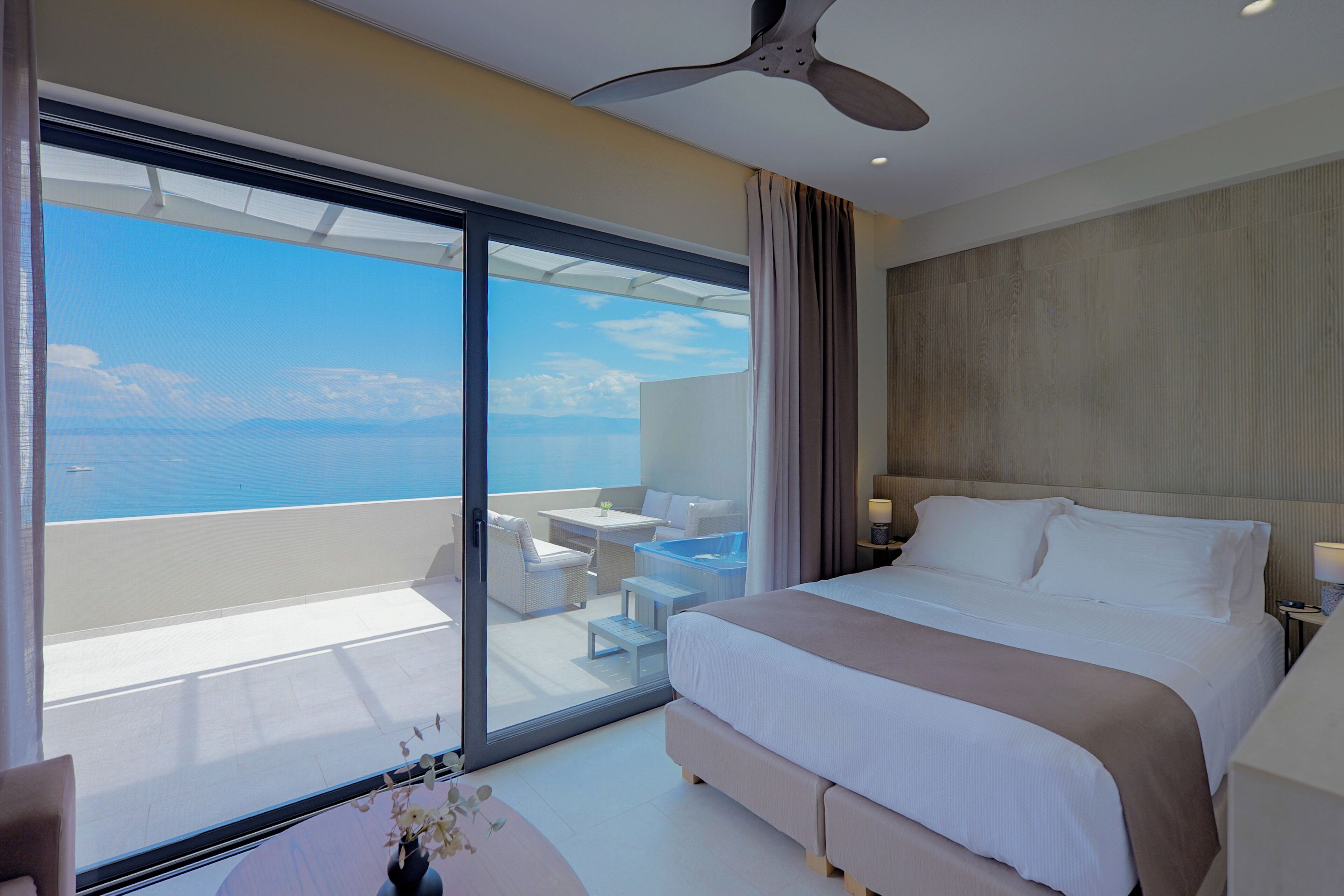 Ein Hotelzimmer mit einem großen Bett, einem Deckenventilator, einem Balkon mit Meerblick und einem kleinen Tisch mit einer Vase.