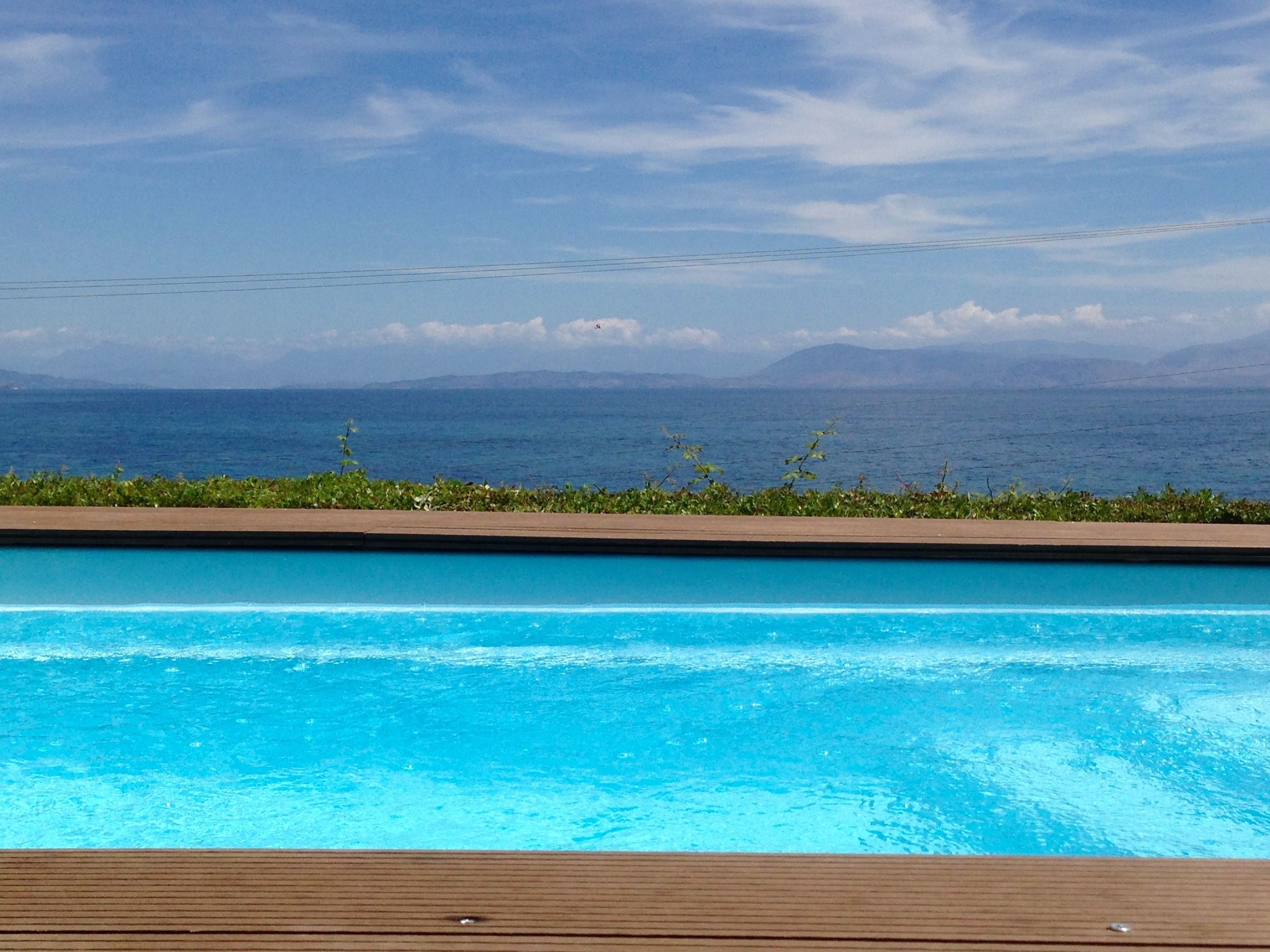A swimming pool with a wooden deck in the foreground and a view of the ocean and mountains in the background.