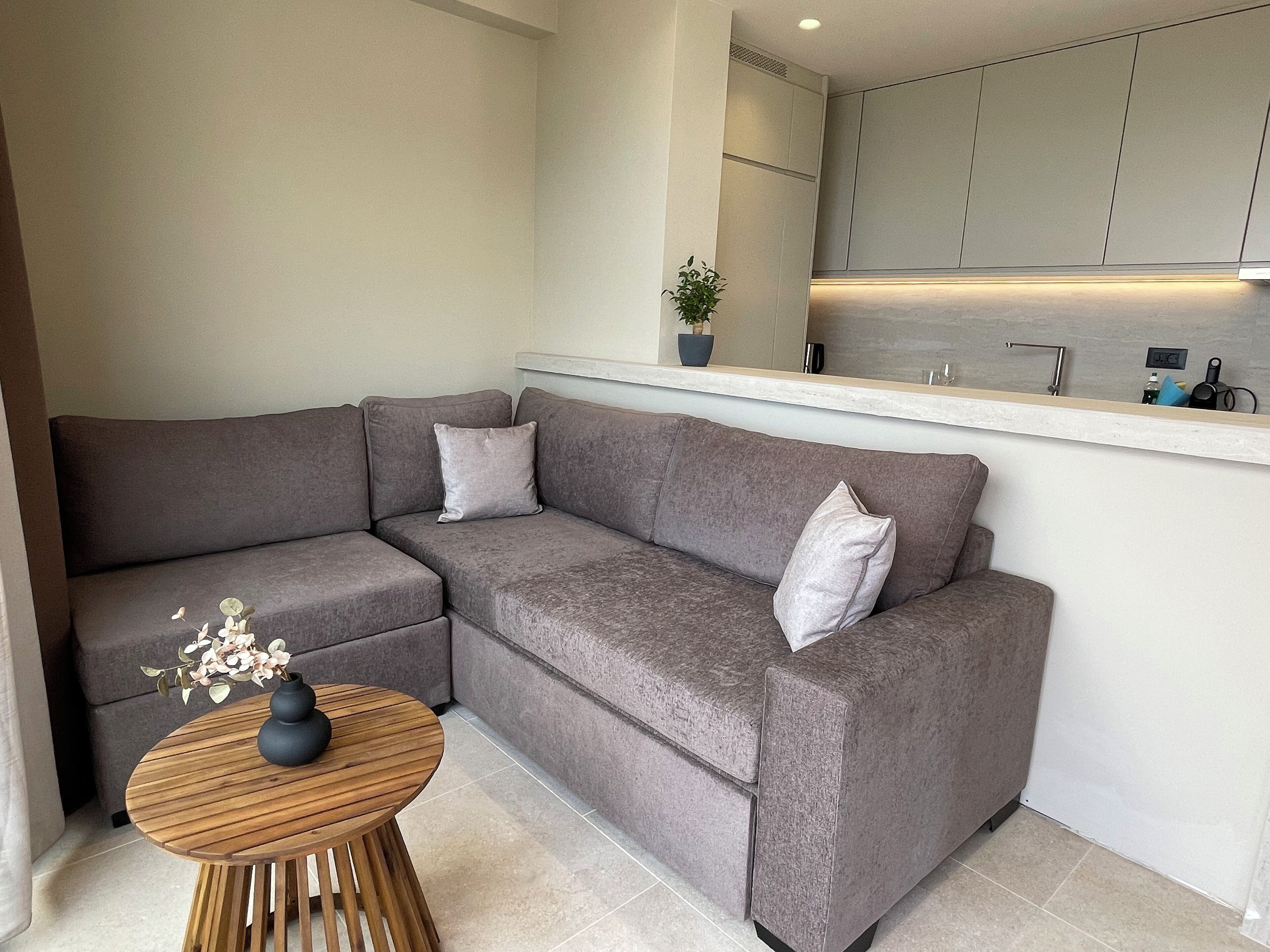 Ein modernes Wohnzimmer mit einer grauen Eckcouch, einem runden Couchtisch aus Holz und einem kleinen Topf mit Pflanze.