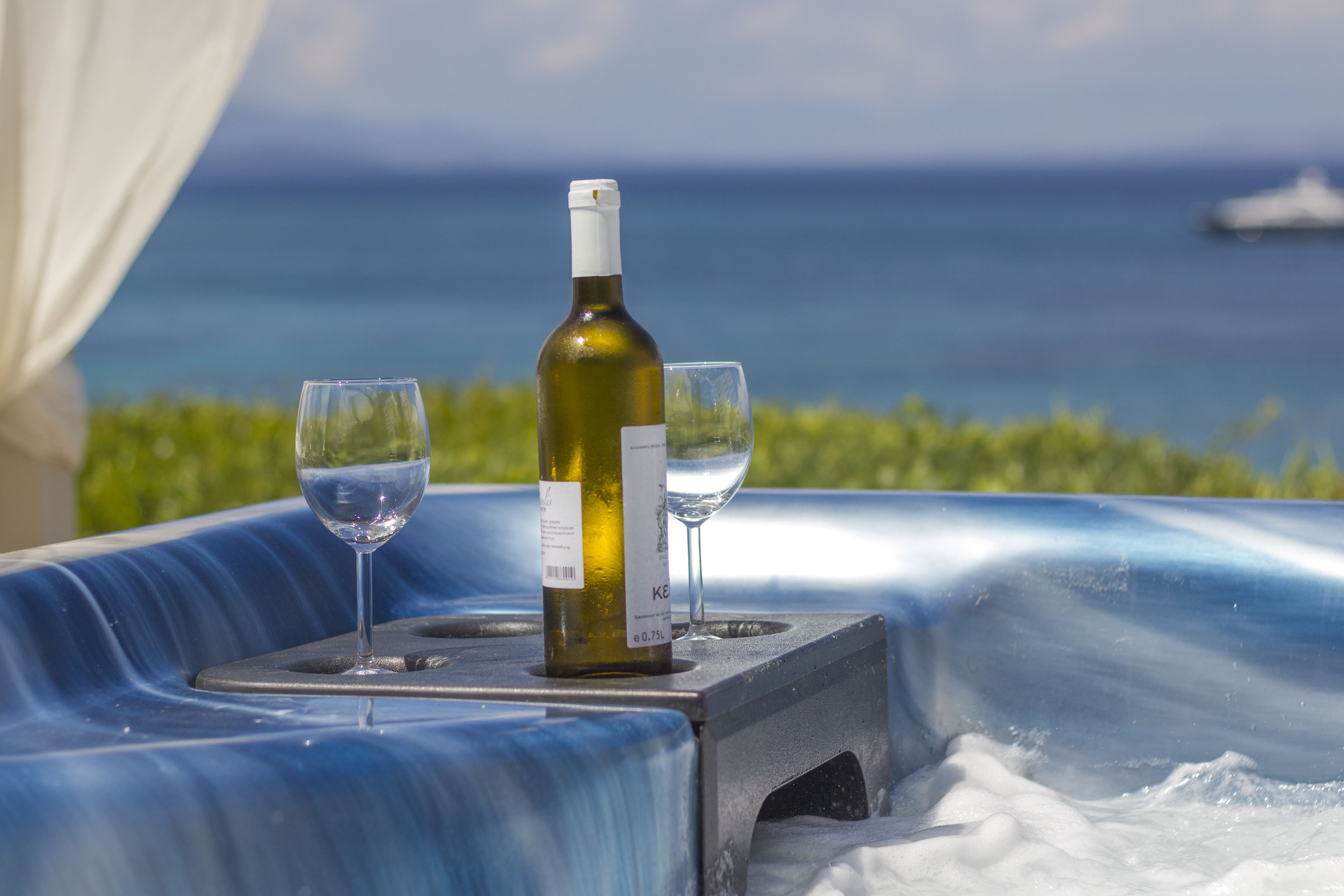 A table with a bottle of wine and two glasses, set against a backdrop of the ocean and greenery.