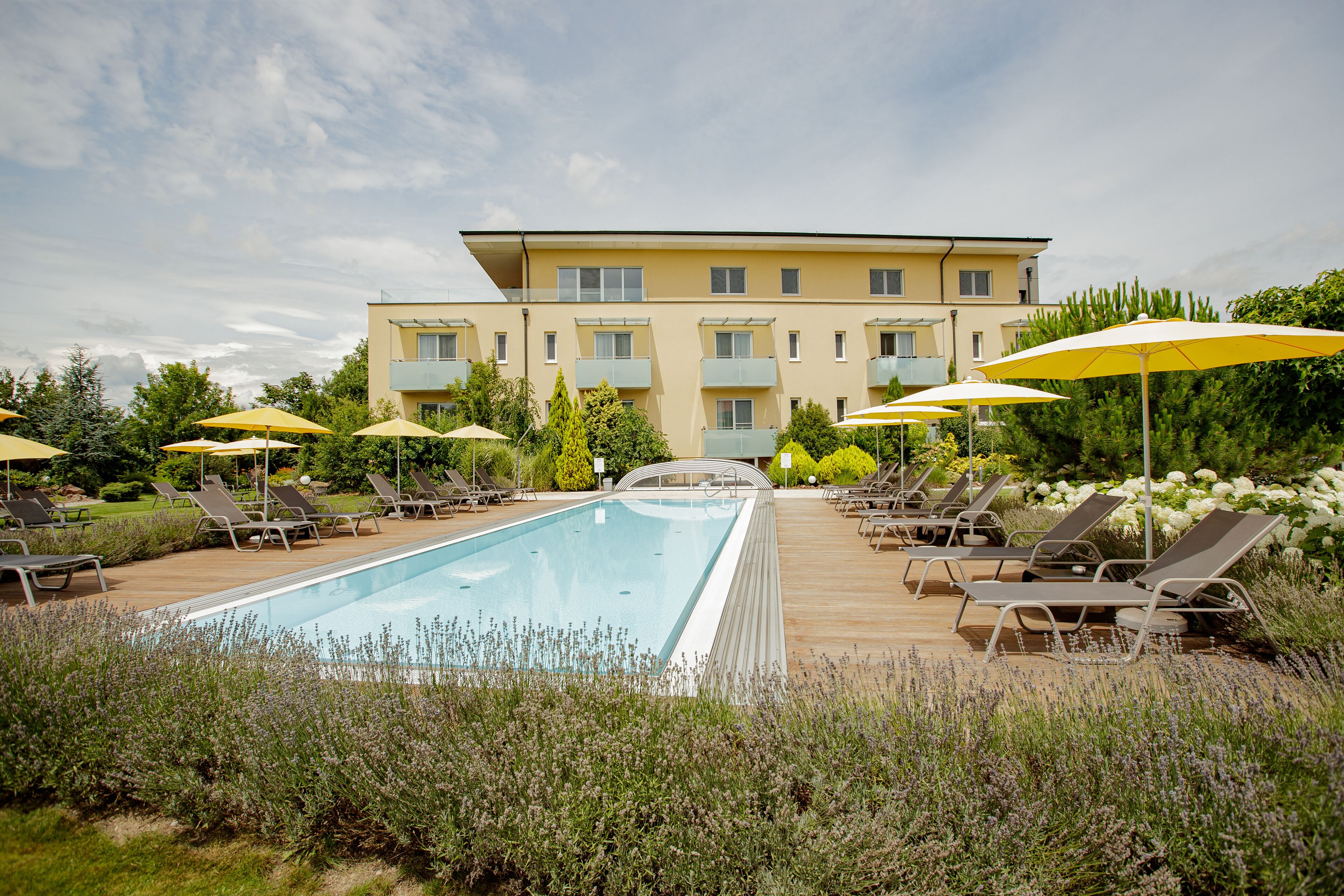 Une piscine entourée de verdure, avec des chaises longues et des parasols, et un immeuble à plusieurs étages en arrière-plan.