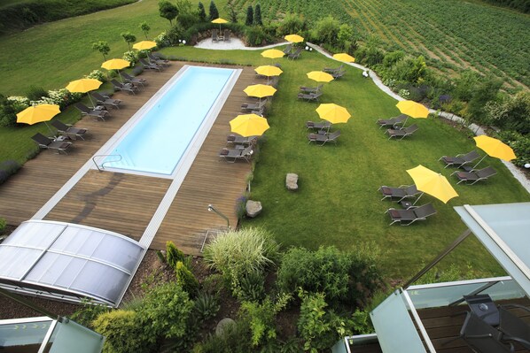 An aerial view of a pool area with sun loungers and umbrellas, surrounded by well-maintained lawns and a wooden deck.