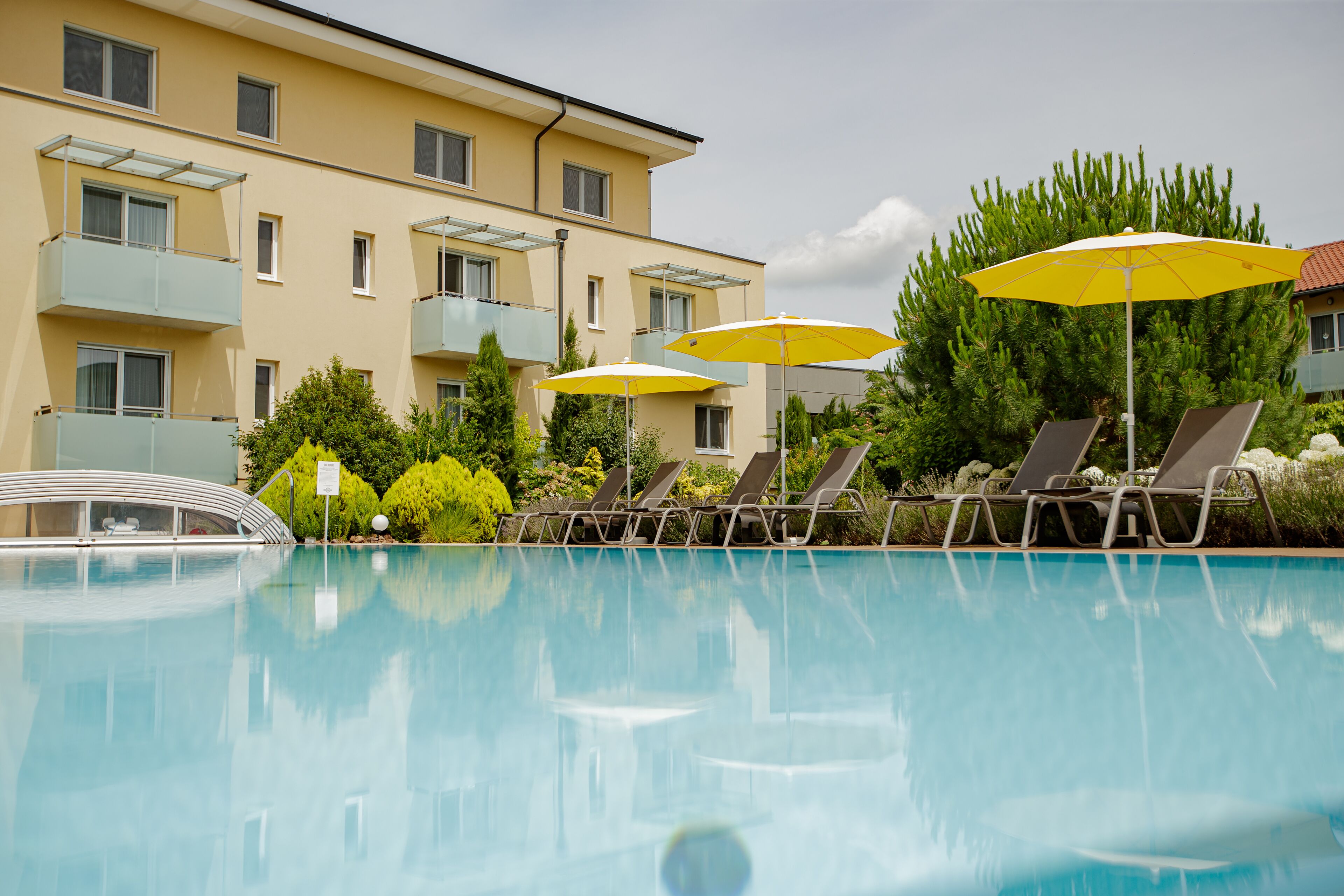 En swimmingpool med liggestole og gule parasoller, omgivet af en bygning med altaner.
