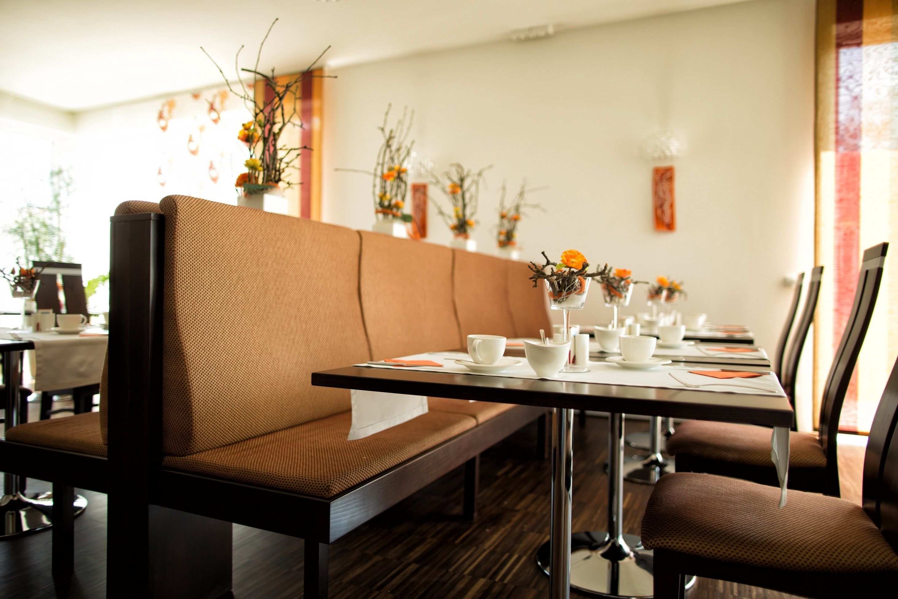 A dining area with a long table, brown upholstered chairs, and a bench. There are flower arrangements on the wall and a painting above the table.