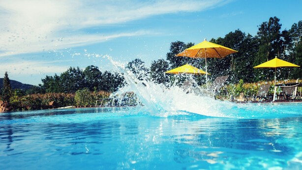 Une piscine d’un bleu éclatant, dont l’eau jaillit en gerbes, entourée de parasols jaunes et de chaises longues.