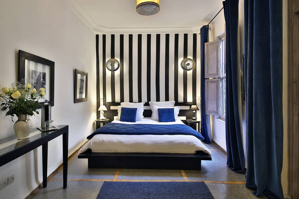 A bedroom with a bed, bedside tables, a nightstand, a wall-mounted light fixture, a window with curtains, and a framed picture on the wall.