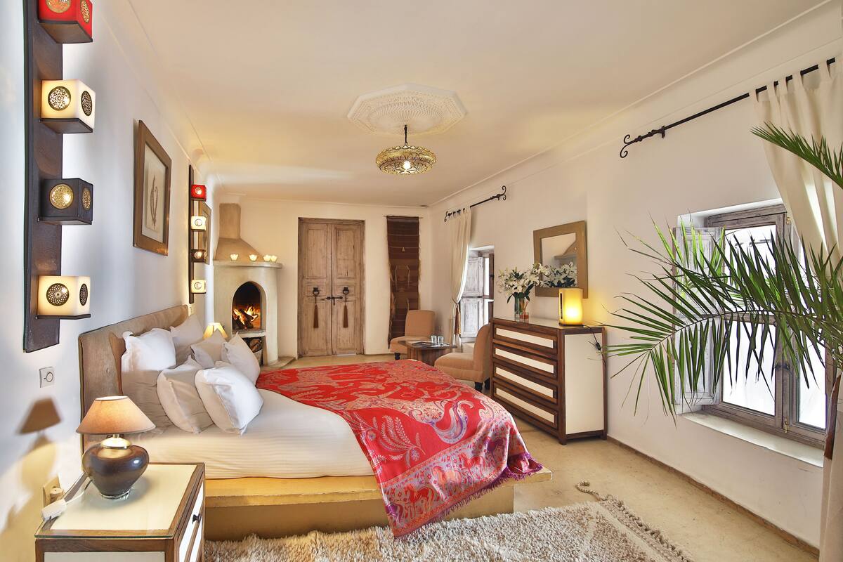 A bedroom with a large bed, bedside tables, a nightstand, a dresser, a mirror, a fireplace, and a window with curtains.
