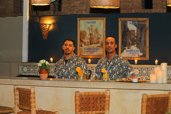 Two baristas in patterned shirts behind a counter with drinks and a vase of flowers. - Riad Anyssates (Marrakech)