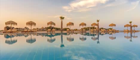 A pool with lounge chairs and thatched umbrellas, reflecting the surrounding palm trees and sky.