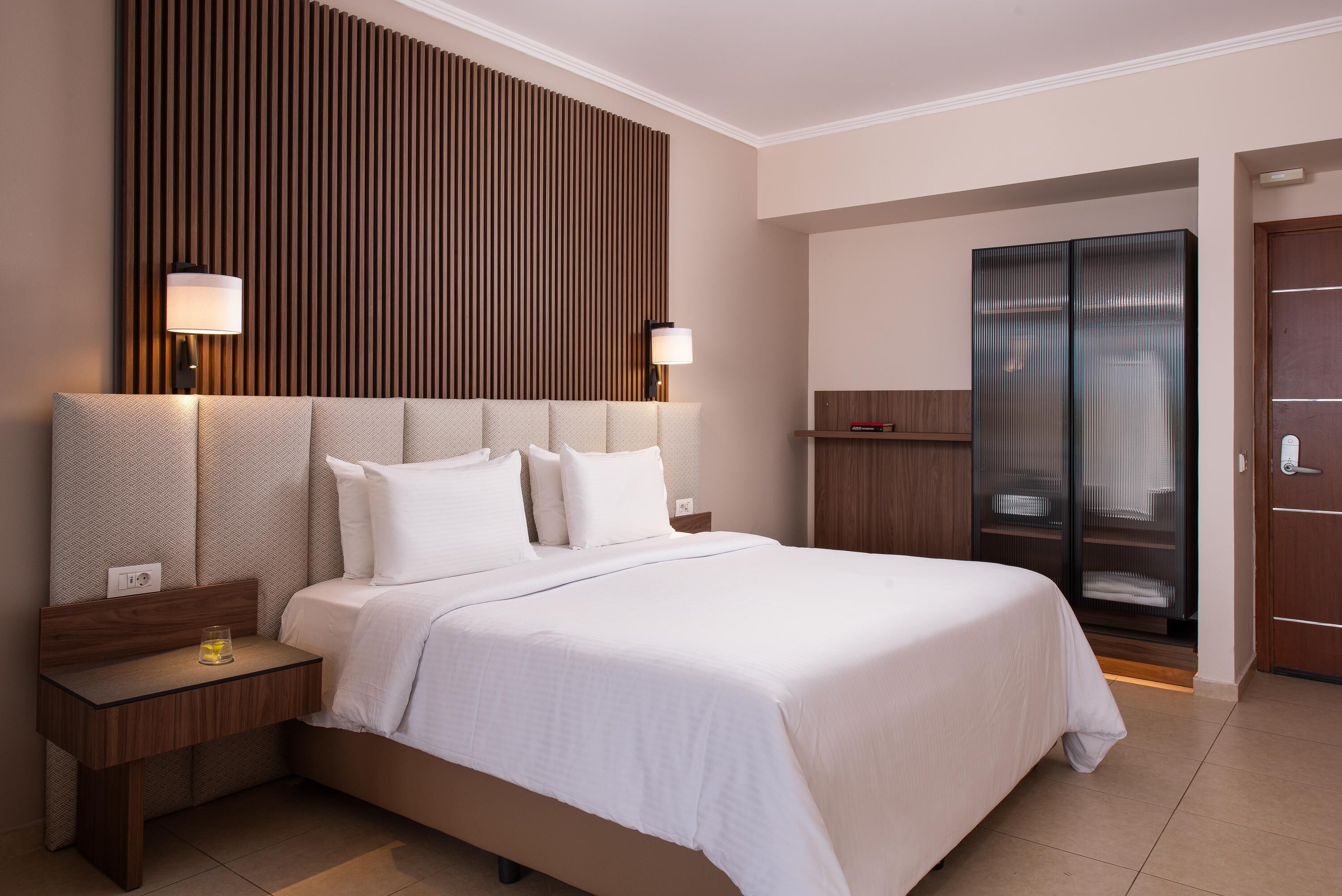 Una habitación de hotel moderna con una cama grande, cabecera de madera, mesita de noche, lámpara y armario.