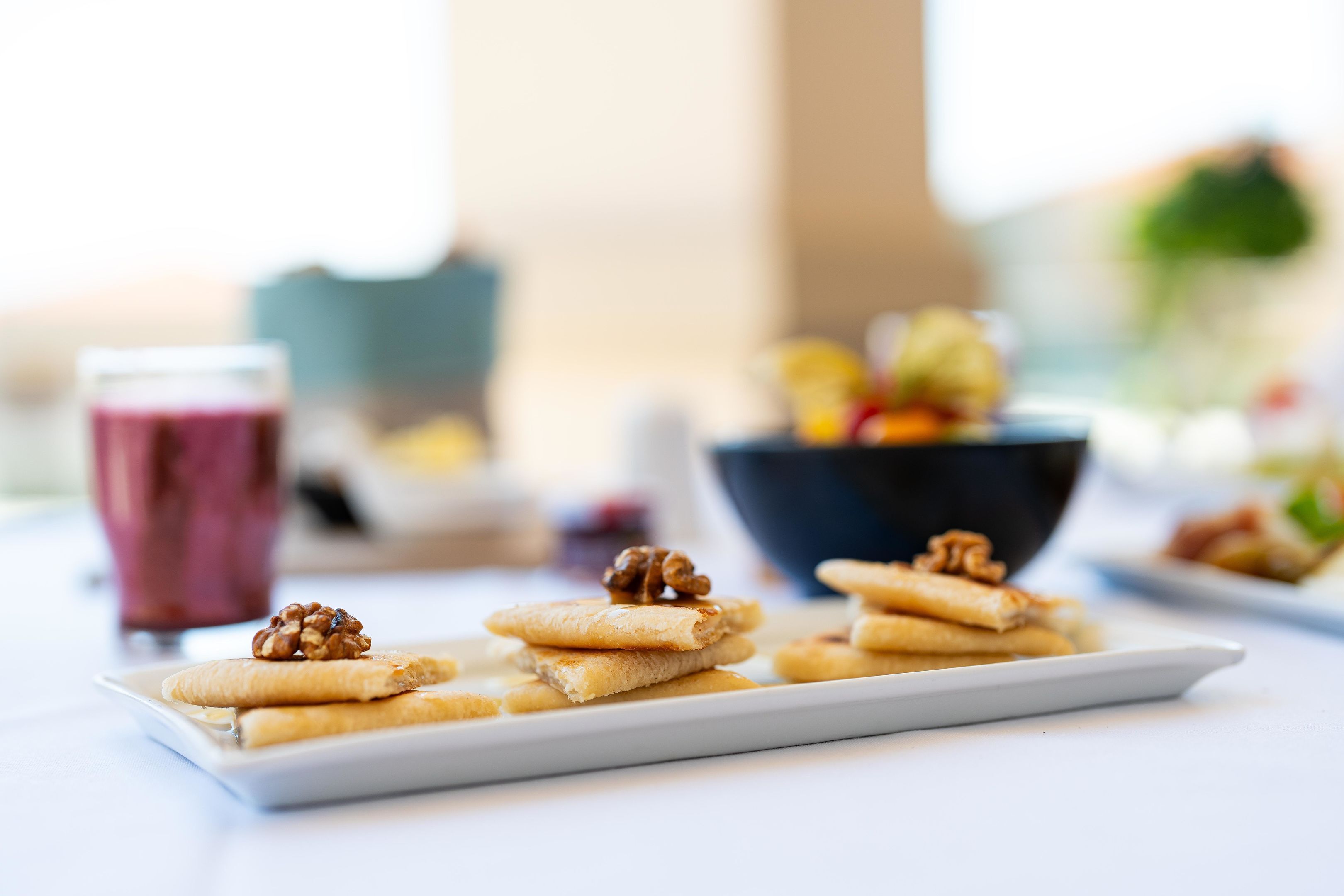 Une assiette blanche sur laquelle sont disposées plusieurs petites pâtisseries garnies de noix, une boisson rose et, au fond, un bol de fruits.