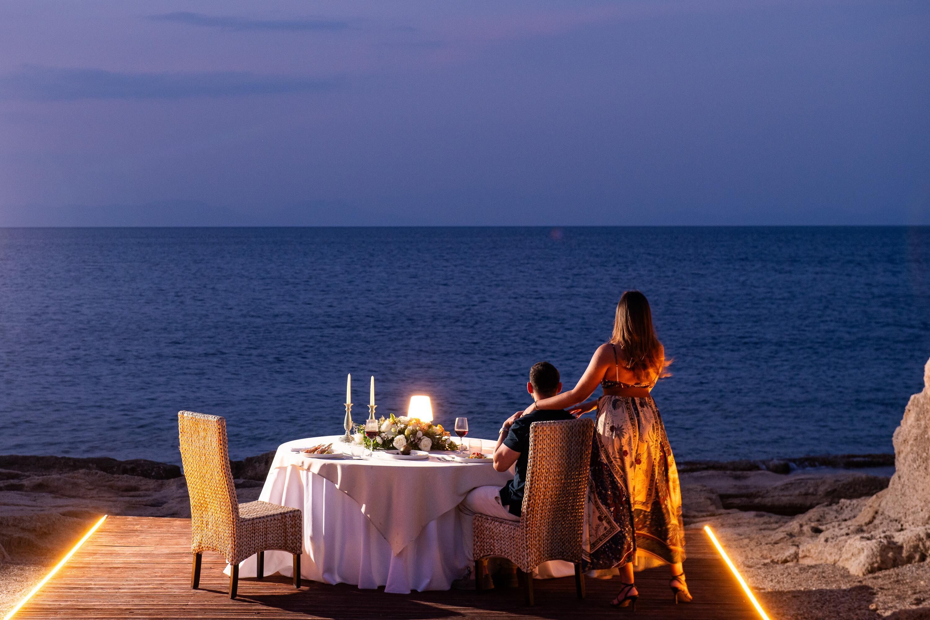 Cena romántica dispuesta en una terraza de madera con mesa para dos, con vistas al mar.