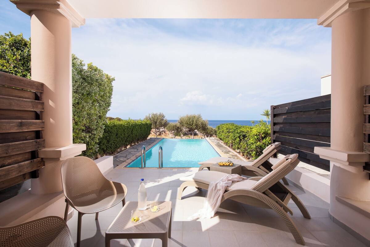 Uma área à beira da piscina com espreguiçadeiras, uma mesa e vista para o mar.