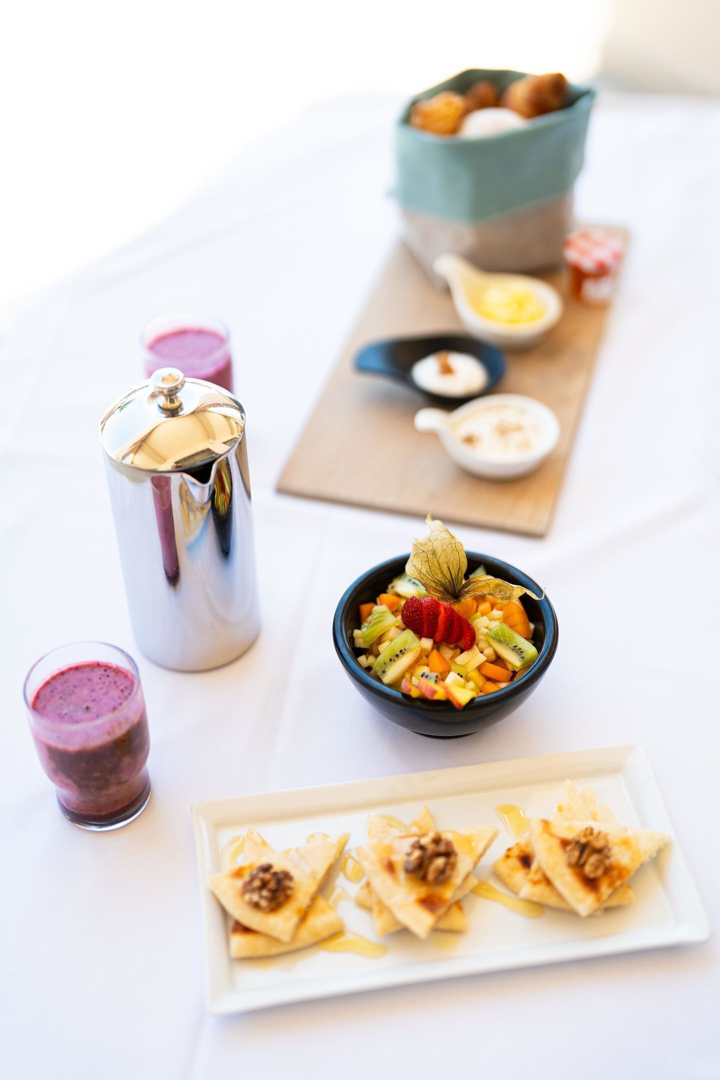 A breakfast setting with a metal coffee pot, a bowl of fruit salad, a plate of pastries, and a cup of smoothie.