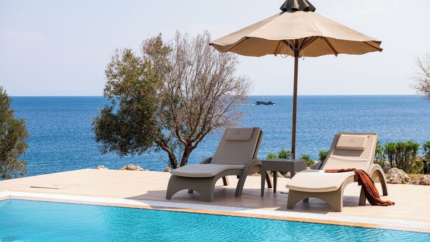 Zona junto a la piscina con sillones de descanso y una sombrilla, con vista a un cuerpo de agua.
