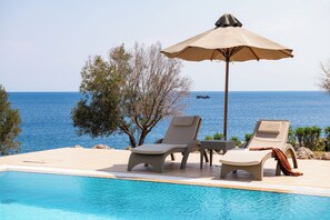 Zona junto a la piscina con sillones de descanso y una sombrilla, con vista a un cuerpo de agua.
