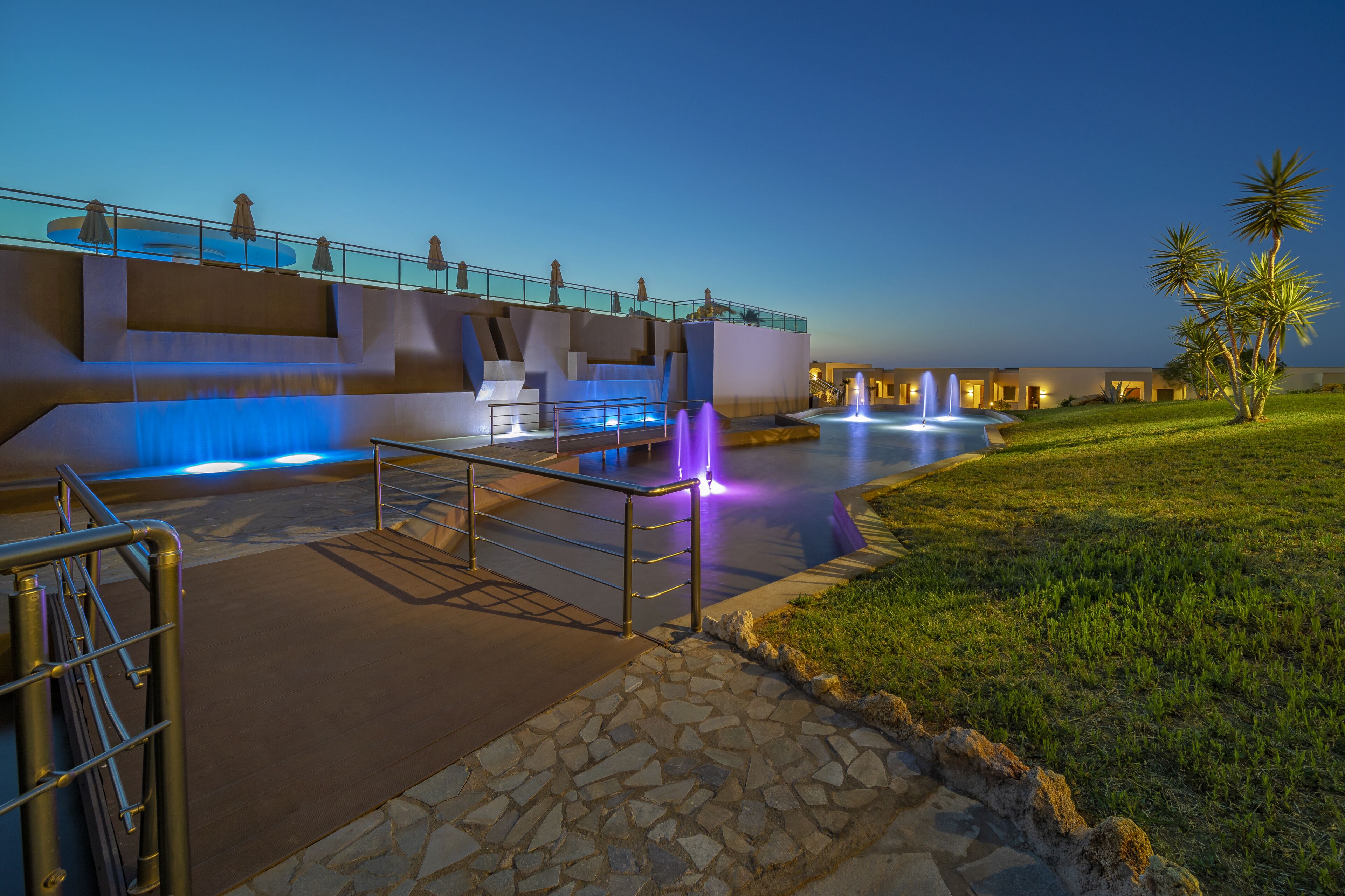 A nighttime view of a hotel with a pool area, outdoor lighting, and palm trees.