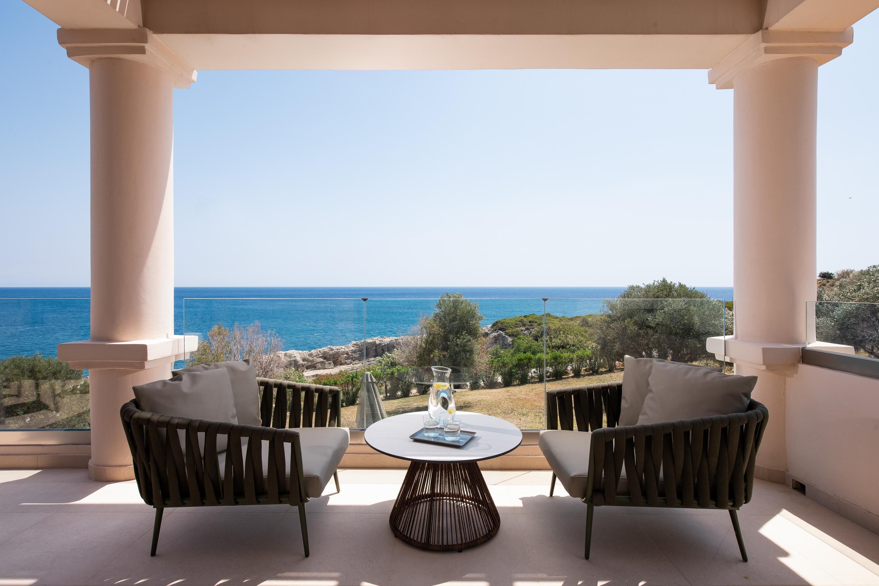 A balcony with two wicker chairs and a small table, overlooking a coastal landscape.