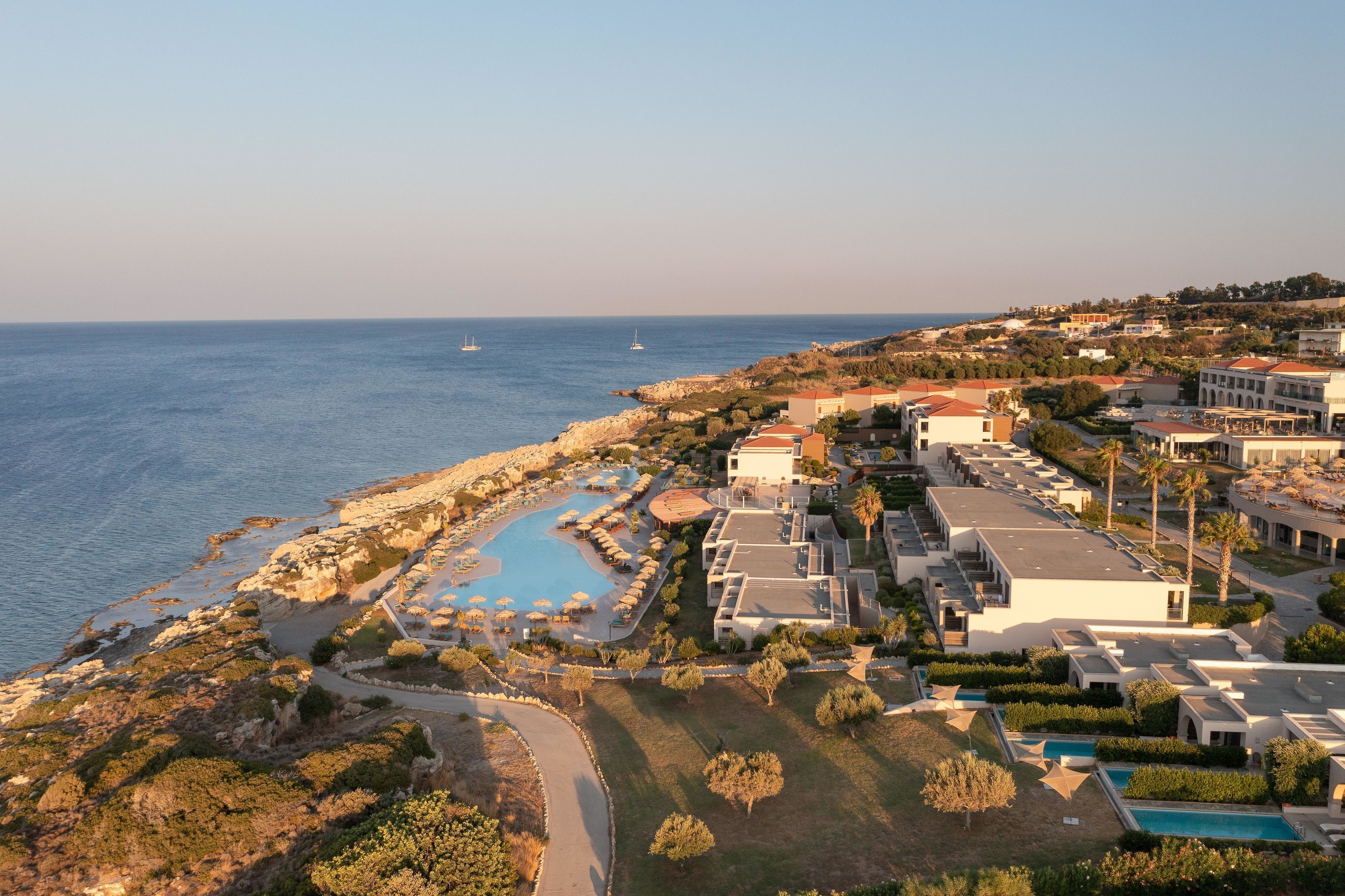 An aerial view of a coastal resort with a swimming pool, multiple buildings, and a view of the sea.