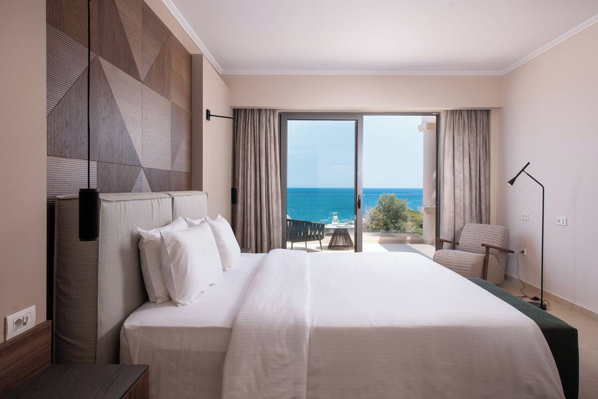 Quarto de hotel com uma cama grande, vista para o mar e uma varanda com mesa e cadeiras.