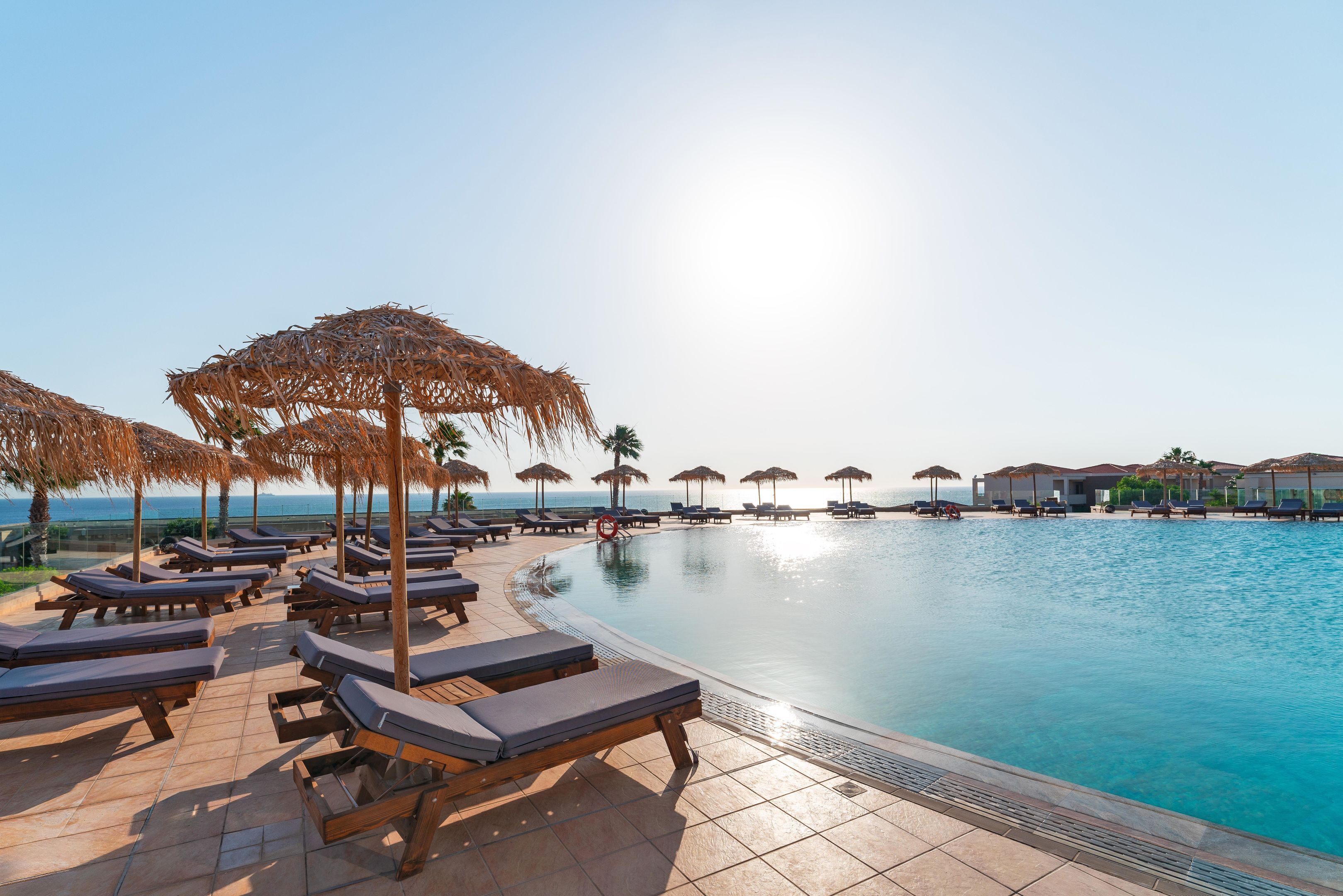 Un espace aménagé au bord de la piscine, avec des chaises longues et des parasols, un ciel dégagé et une vue sur la mer.