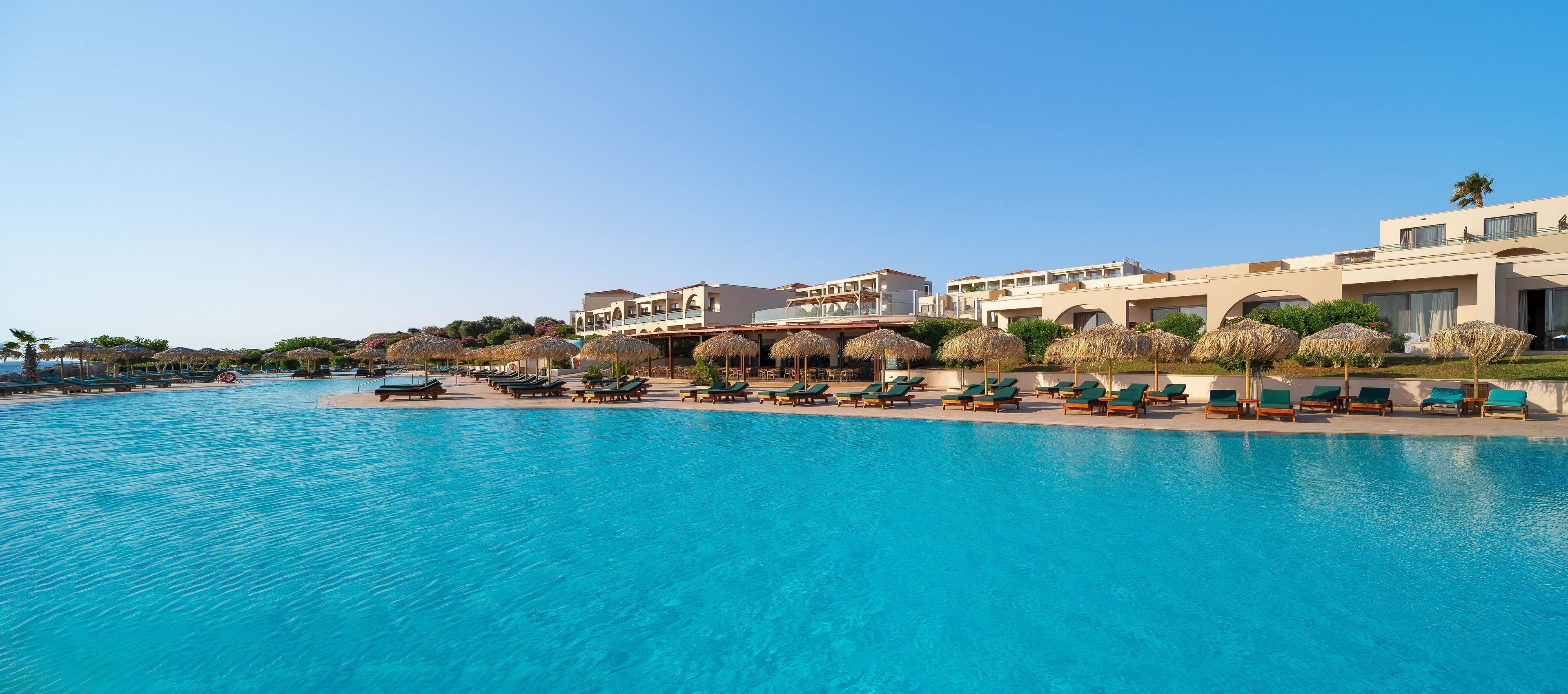 A large swimming pool with lounge chairs and umbrellas, surrounded by buildings with balconies and a clear sky.