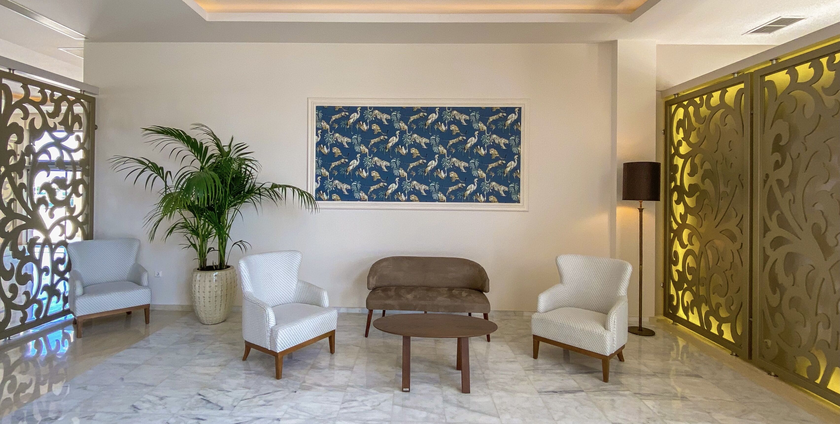 A lobby with a marble floor, two white armchairs, a brown sofa, a large potted plant, a framed tropical pattern artwork, and decorative panels.