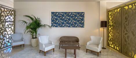 A lobby with a marble floor, two white armchairs, a brown sofa, a large potted plant, a framed tropical pattern artwork, and decorative panels.
